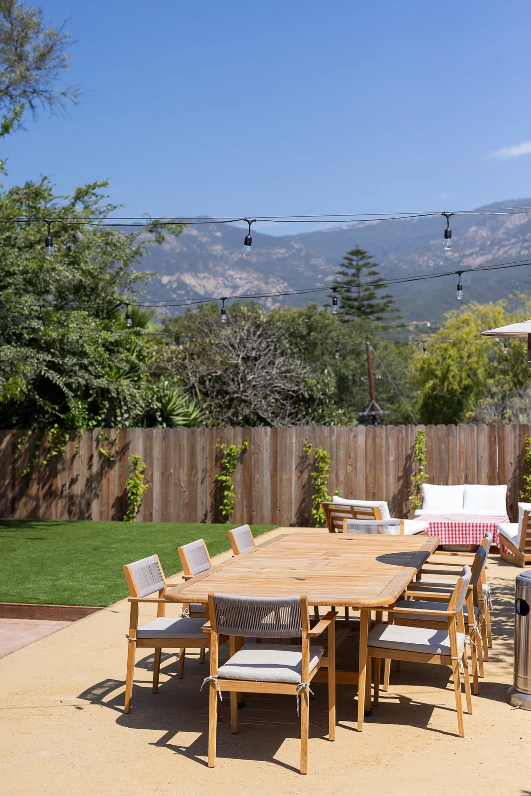 Long teak dining table under string lights with the mountains beyond